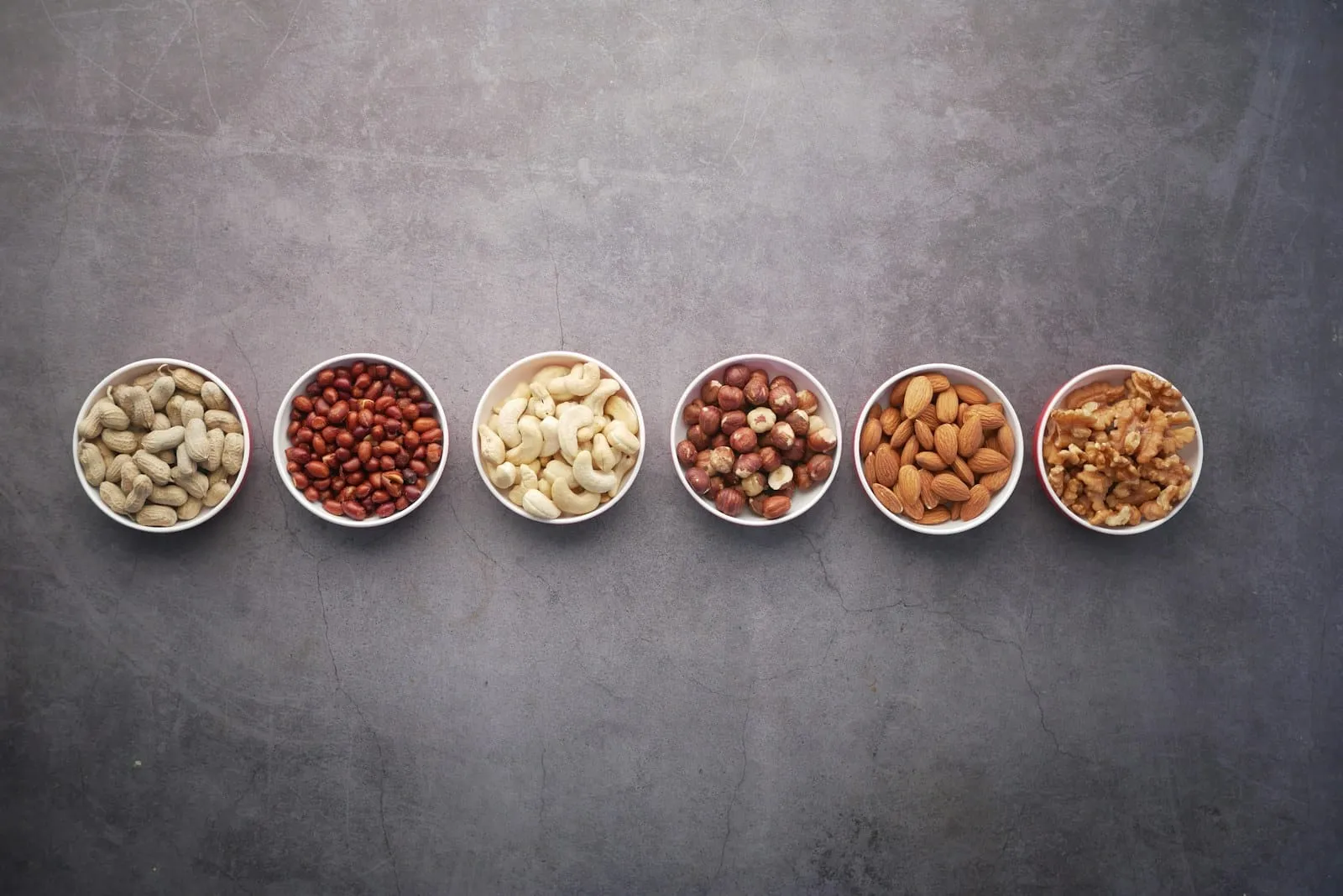 Six small bowls of nuts and seeds lined up on a slate benchtop.