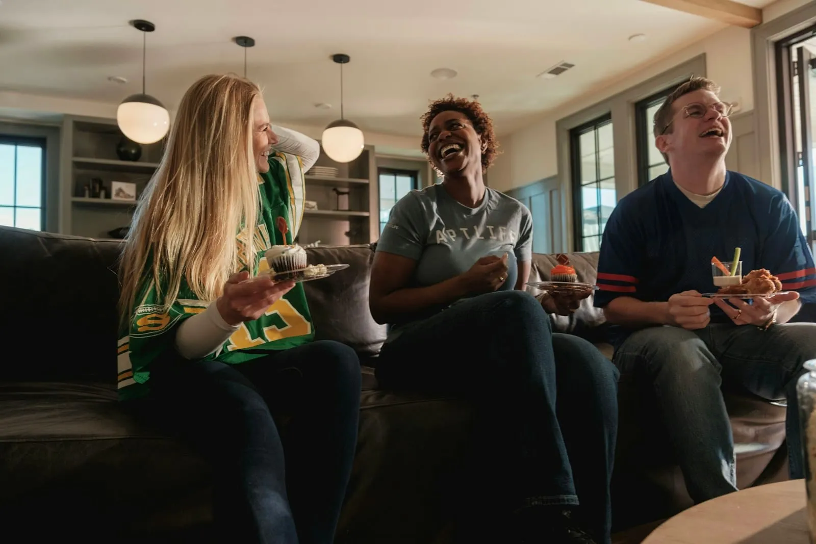 Three friends laughing together on a couch with plates of food.